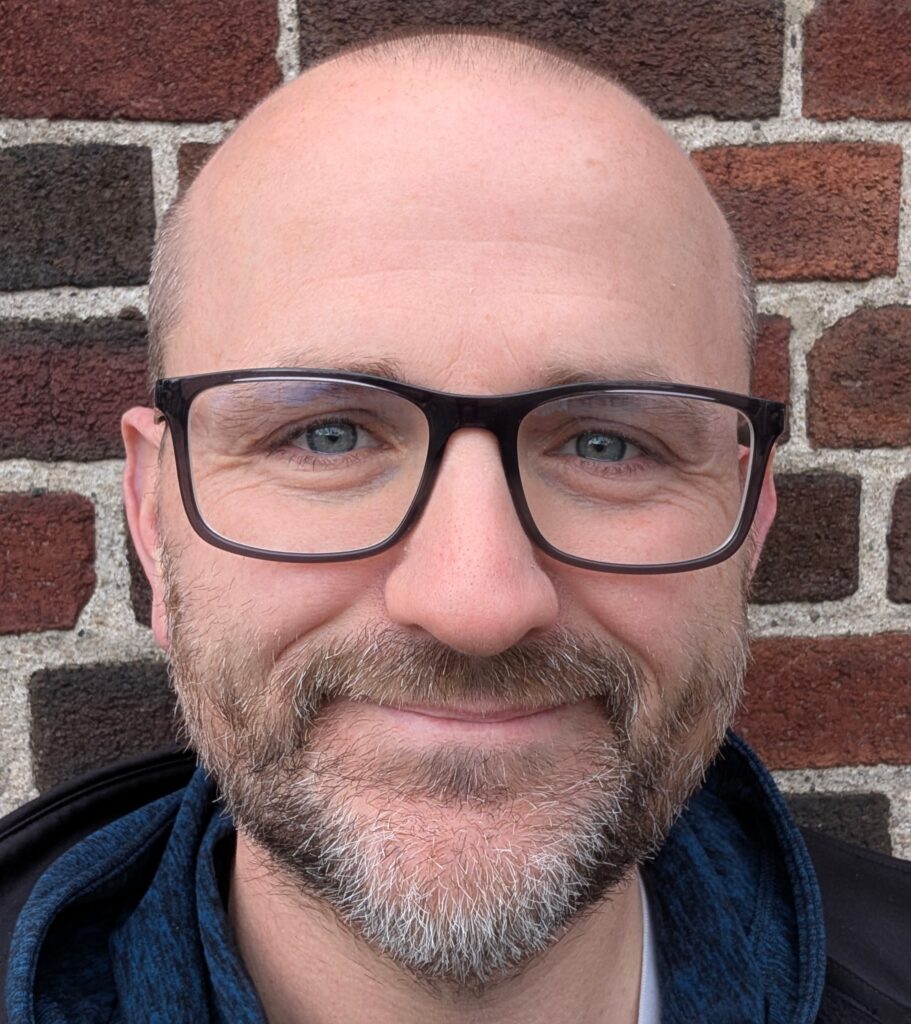 Headshot of Lance Eaton smiling while standing against a brick wall
